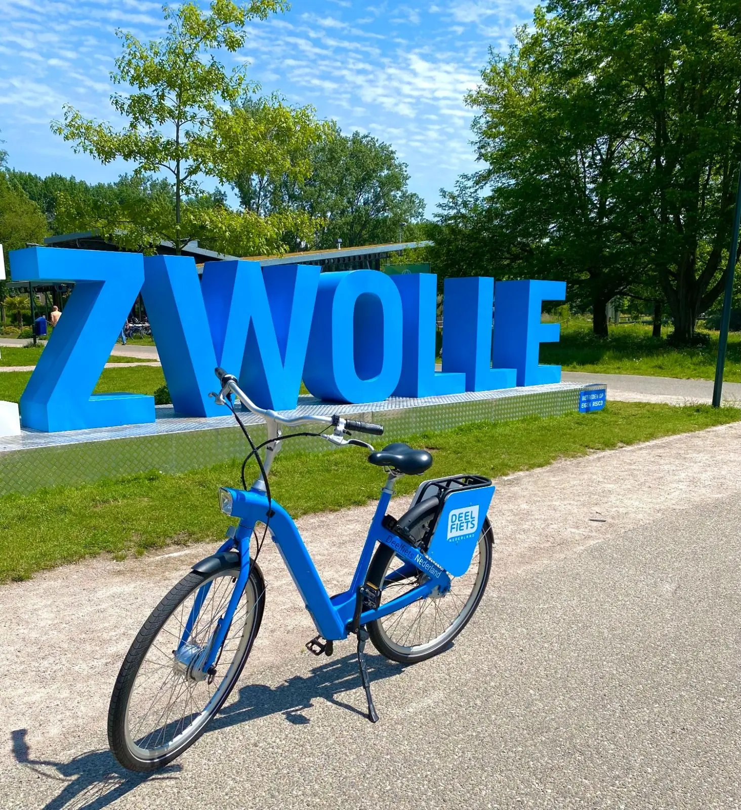 Een deelfiets van Deelfiets Nederland met een groot, blauw bord in de achtergrond met de tekst Zwolle.