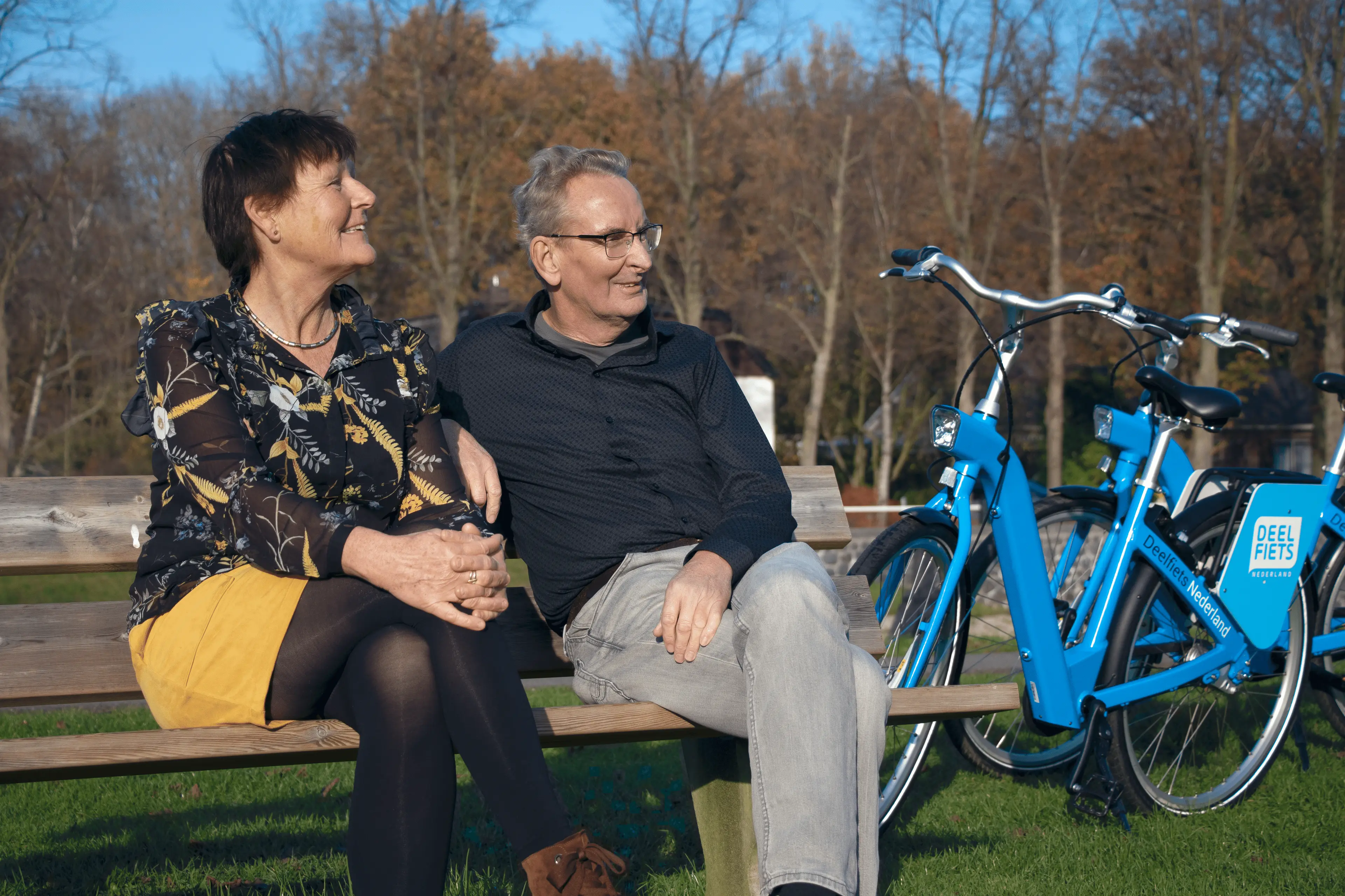 Twee blije gebruikers van de Deelfiets die rusten op een bankje.